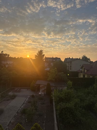 This is a high-angle, medium shot capturing a serene sunset in Marki, Poland. The sun, a vibrant orange orb, is positioned in the upper left quadrant of the frame, casting a warm glow across the sky and illuminating a scattering of textured clouds. The sky transitions from a bright yellow and orange near the sun to a soft blue at the top of the image.
Below the sky, a dense line of trees forms a natural barrier. Behind the trees, several buildings are visible, including residential houses with red-tiled roofs and apartment buildings with flat roofs. One prominent house in the mid-right of the frame features a yellow facade and a dark red roof.
The foreground depicts a backyard area. A paved pathway, made of rectangular stones, leads diagonally from the lower left towards the center. To the left of the pathway, a fenced area contains a wooden picnic table with benches. In the center of the frame, a small, shed-like structure with a brick base and a dark roof stands amongst lush green foliage. The ground in the foreground is a mix of dark soil, scattered stones, and low-lying green plants. The overall impression is one of a tranquil, residential evening in a suburban setting.