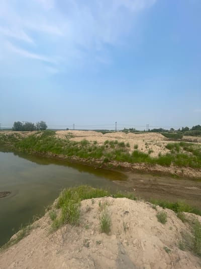 An outdoor, eye-level, wide shot captures a barren landscape of sand dunes next to a body of muddy water under a hazy, pale blue sky. The foreground is dominated by a steep, sandy embankment covered in sparse green grass. To the left, the murky water reflects the muted sky. In the middle ground, more sand dunes rise, interspersed with patches of green grass and vegetation. Beyond these dunes, a flat expanse of land is visible, with distant trees and several tall, thin power pylons supporting overhead electrical wires. The overall impression is one of a construction or excavation site, possibly in the outskirts of Yermolayevo, Russia, given the presence of industrial infrastructure. No people or animals are visible.