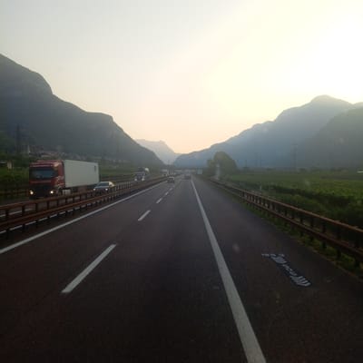 The image captures a perspective from a vehicle traveling on a highway in Brentino, Italy, during what appears to be late afternoon or early evening, given the soft, diffused light. The highway stretches into the distance, flanked by steep, verdant mountains on either side. The road is paved and marked with white lane lines. Several vehicles are visible on the highway, including trucks and cars. A prominent red and white truck, identified by the lettering "BROGIE LOGISTICS" on its side, is in the left lane, moving forward. Further ahead, other trucks and cars are also traveling. On the right side of the highway, there is a vineyard with rows of grapevines. The sky is a pale, hazy yellow, indicative of either sunrise or sunset, or possibly atmospheric haze. The overall scene depicts a typical highway transit through a mountainous, agricultural region.