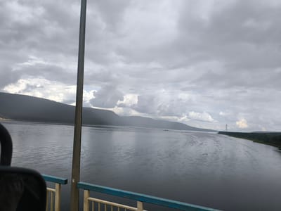 The image captures a wide, calm river under a heavily overcast sky. The water is dark and reflects the grey clouds, with subtle ripples on its surface. On the left bank, a dark, treed hillside stretches into the distance, fading into a hazy background. A tall, thin metal tower stands on the right bank, silhouetted against the sky. In the foreground, the edge of a railing with blue and yellow metal bars is visible, suggesting the viewpoint is from a bridge or elevated platform. A dark, curved object, possibly part of a seat or structure, is in the lower left corner. The overall mood is somber and tranquil. This scene is likely near Angarsk, Russia, given the characteristic landscape of the region featuring large rivers and surrounding forested hills.