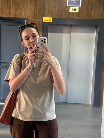 A young woman is taking a mirror selfie in what appears to be an elevator. She is wearing a light-colored t-shirt, dark brown pants, and sunglasses on her head. She is holding her smartphone in her right hand, which is angled to capture her reflection. Her left hand is holding a set of keys and displaying several rings on her fingers. A small tattoo of a spade can be seen on her left forearm. She has dark hair and appears to be smiling faintly. A brown handbag is slung over her left arm.
The elevator has a grey, metallic door and paneling. The wall to the left of the elevator appears to be wood-paneled, with a small yellow sign attached. There is a blue-framed sign on the wood paneling with some text on it, and a small light with green illuminated numbers or symbols. The floor is tiled with a grey, speckled pattern. The overall impression is a contemporary, indoor setting, likely in a residential building or a public space like a hotel or office. The lighting suggests it is daytime, as there is ambient light reflecting off the elevator doors. There is no clear indication of the specific time of day or weather conditions outside.