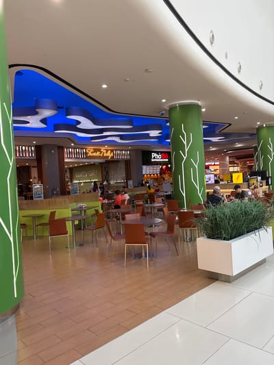 The image depicts a food court area within a shopping mall, featuring a vibrant and modern design. Large, green, cylindrical pillars with white tree-like graphics are a prominent element of the interior architecture. The ceiling is adorned with a blue, wave-like design, creating a dynamic visual effect.
Several food stalls are visible, including "PhoBo" and "Naked Chef," suggesting a variety of culinary options. Tables and chairs, predominantly in brown and orange hues, are arranged throughout the space, with some occupied by patrons. The flooring is a mix of wood-look tiles and white tiles. A white planter box filled with lush green foliage is placed in the foreground, adding a touch of nature to the urban setting. The overall atmosphere appears to be casual and bustling, typical of a food court environment. The location of Tlustenkhabl, Russia, is noted as the setting, though no specific landmarks or identifiers within the image confirm this directly.