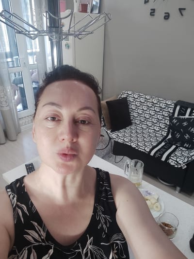 The image shows a close-up selfie of a woman with medium-length dark hair, wearing a black sleeveless top with a floral pattern. Her head is tilted slightly to the side, and she is looking at the camera with her lips pursed. In the background, a living room is visible, with a sofa covered in a black and white patterned fabric that appears to be designer logos. There are several pillows on the sofa, some plain and some with the same logo pattern. A blanket with similar patterns is draped over the back of the sofa. To the left of the woman, a white table is visible, with a remote control, a glass mug, a small plate with sliced bananas, and a small glass with a brown liquid. The room has light-colored walls and a large window with sheer curtains. The overall setting suggests an indoor environment, likely a home. There is no indication of the city of Minglola mahalla, Uzbekistan.