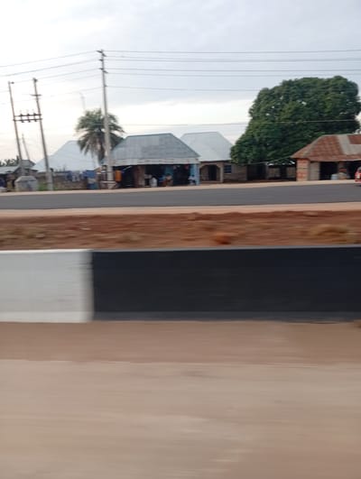 The image depicts a roadside scene in Uke, Nigeria, captured from a moving vehicle. The foreground consists of a blurred, light brown-tan surface, followed by a black and white concrete barrier (white on the left, black on the right) exhibiting motion blur. Above the barrier, a strip of reddish-brown earth indicates a roadside verge.
The midground features a dark grey asphalt road, appearing recently paved. Along the opposite side of the road, a series of single-story structures are visible. On the left, utility poles with multiple power lines extend across the frame, partially obscuring distant residential buildings. Further right, a cluster of at least three buildings with grey corrugated metal roofs are present; one appears to be an open-front shop. Behind these, a tall palm tree is visible. Towards the right, a large tree with dense green foliage stands prominently. Adjacent to it, another building with a rusted brown corrugated roof, resembling a shop, is visible. A red vehicle is partially discernible on the far right, near this last structure.
The background is dominated by an overcast sky, uniformly light grey with cloud cover. No individuals are clearly identifiable due to motion blur, though indistinct shapes near the shops suggest potential human presence. The scene collectively represents a semi-urban or rural setting characterized by local architecture and recent infrastructure development.