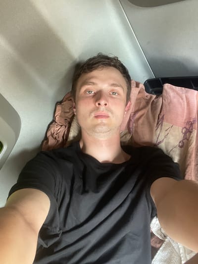 A man is lying on his back, with his head resting on a pink and beige patterned blanket. He is wearing a black t-shirt. The man has light skin, short brown hair, and green eyes. He is looking directly at the camera with a neutral expression. His right arm is extended towards the camera, as if he is taking the photo himself. The background appears to be a neutral colored surface, possibly a bed or a couch, with a dark object behind the blanket and to the left of the man's head. There is a white crocheted item to the right of the man's head, partially covered by his arm. The lighting suggests an indoor setting, likely during the day. No visible text is present in the image.