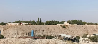 The image depicts an industrial site, likely a quarry or construction material processing facility, located in Margilan City, Uzbekistan. The landscape is dominated by a large, excavated earth embankment, revealing layers of soil and rock. Several structures are visible, including industrial buildings with metal roofs, storage silos (one distinctly blue), and smaller sheds. Conveyor belts and metal frameworks suggest ongoing material handling and processing. A few residential or administrative buildings with simple designs are also present, situated on the flatter ground above the embankment. The background is characterized by a sparse line of trees and vegetation, with power lines and transmission towers indicating the presence of electricity infrastructure. The sky is clear and light blue, suggesting a daytime scene. The ground in the foreground is covered with loose rocks and debris, consistent with a quarry or construction site. No people are visible in the image.