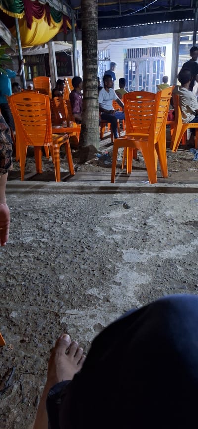 In Payaterbang, Indonesia, a group of people are gathered outdoors, possibly at a food stall or event. The scene is lit by artificial lighting, creating a warm and somewhat hazy atmosphere. Bright orange plastic chairs are scattered throughout the area, some occupied by individuals. In the foreground, a person's bare foot rests on the textured ground. Behind the chairs, a thatched roof structure is visible, adorned with colorful fabric drapes in shades of yellow, red, and green. A tall, slender palm tree trunk stands prominently in the mid-ground, partially obscuring the view of some of the people. Several individuals are seated, appearing engaged in conversation or observation. The ground is uneven, with patches of gravel and what looks like hardened dirt or concrete. The background shows a building with doors and windows, suggesting a more permanent structure behind the outdoor setup. The overall impression is of a casual gathering or social space in a tropical environment.