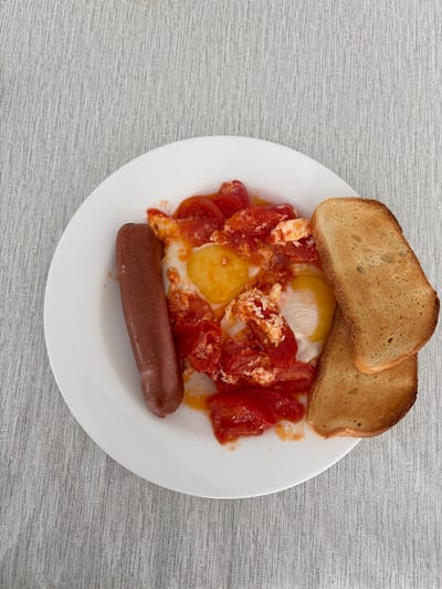 A white plate contains a breakfast meal. On the left side of the plate is a single, reddish-brown sausage. In the center of the plate, there are two fried eggs mixed with chopped tomatoes. To the right of the eggs are two slices of toasted bread, stacked slightly. The plate rests on a grey, textured surface, likely a tablecloth. The scene is photographed from a top-down perspective. The location is not discernible from the image itself.