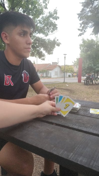 A person in a black shirt with a red logo is looking at the camera, sitting at a wooden picnic table outdoors. Someone else's arm and hands are also visible, holding a hand of Uno cards. A stack of cards sits on the table next to the hands. In the background are trees, a street with a building, and two bicycles leaning against a post. The sky is overcast.