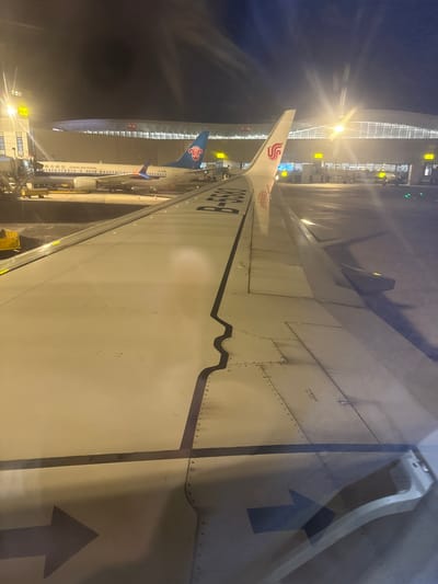 The image shows a section of an airplane wing, likely taken from the interior of the plane near the window, as the camera's vantage point is looking out. The wing is predominantly white with visible rivets along its surface, and a black line runs along its length. There's an arrow graphic painted on the wing. Visible through the window is an airport scene at night, with bright lights illuminating the jet bridge and another plane, presumably a China Southern Airlines aircraft, parked nearby. The aircraft tail features the China Southern logo. The tail number of the adjacent plane is partially visible, appearing to read "B-2206". The location is Ürümqi, China. No people are directly visible in the frame.