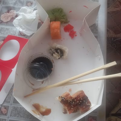 In Cepeleuți, Moldova, this image captures the aftermath of a meal within a white, folded paper takeout box. The box contains remnants of sushi, including a single piece of orange-colored sushi, a small mound of green wasabi, and two pieces of sushi rolls topped with sesame seeds and a glaze. A small plastic container with dark soy sauce is also present. Chopsticks rest diagonally across the box, implying they were used for eating. Scattered around the box are small pieces of what appear to be pink pickled ginger and a crumpled white tissue. The box itself is placed on a surface with a grey and white floral patterned tablecloth. To the left of the box, a red cardboard insert with two circular cutouts is visible. The overall scene suggests a meal that has recently concluded, with the disposable packaging indicating a casual dining experience.