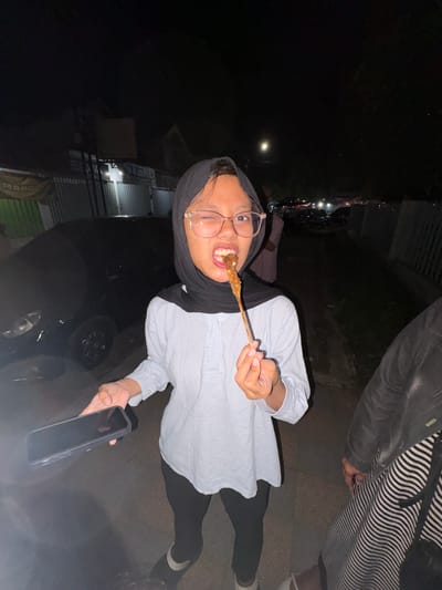 In Tanggul, Indonesia, at night, a young woman wearing a black hijab and glasses is pictured. She is dressed in a light blue and white striped long-sleeved shirt and black leggings. She is holding a smartphone in her left hand and eating a piece of food on a skewer with her right hand, bringing it to her mouth. She is standing outdoors, with a dark-colored car visible behind her. In the background, there are streetlights and signs that suggest a marketplace or food stall area. Another person, partially visible on the right, is wearing a black jacket over a black and white striped garment. The overall lighting is dim, characteristic of nighttime.