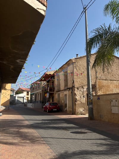 In Monteagudo, Spain, a narrow street is lined with buildings decorated with colorful triangular flags strung across the sky. The street is paved with cobblestones in shades of red and gray, with shadows cast by palm trees. A red car is parked on the side of the street in front of a tan building. The buildings appear old and weathered, with some showing signs of disrepair. The sky is clear and blue.