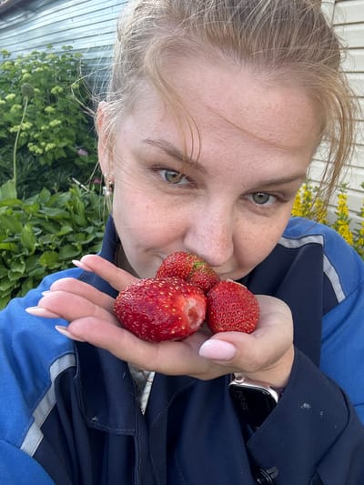 The image shows a close-up, selfie-style shot of a young woman holding three strawberries in her left hand. She is wearing a dark blue jacket with light blue accents. Her hair is light blonde and appears to be tied back loosely, with some strands framing her face. She has green eyes and fair skin with freckles. The strawberries are bright red and appear to be fresh. In the background, there is lush green foliage and a portion of a light-colored, corrugated building, suggestive of a rural or suburban setting. The lighting suggests it is daytime. The location is specified as the city of Dunilovo, Russia.