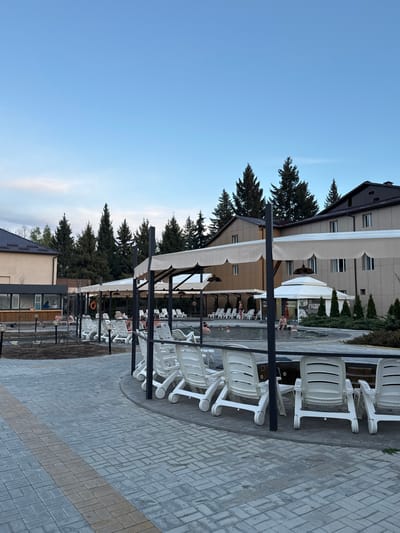 The image shows an outdoor scene in Kara-Oy, Kyrgyzstan, featuring a swimming pool area. Rows of white plastic lounge chairs under awnings are arranged along a paved walkway adjacent to the pool. The pavement is made of grey interlocking bricks, with a band of tan bricks running along one side. Trees and buildings are visible in the background. People are in the pool.