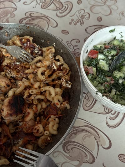 A top-down shot depicts a meal on a patterned tablecloth, possibly taken indoors. In the upper left, a white bowl contains a salad with cucumbers, tomatoes, and greens. Below, a frying pan holds cooked pasta, possibly elbow macaroni, with fried meat and onions. Two forks are visible, one embedded in the pasta and the other alongside the pan. The tablecloth displays a coffee bean and coffee cup design. The location is likely a private residence, not specifically identifiable as Uvarovo, Russia without additional context.