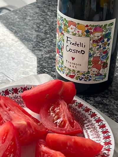 A close-up shot captures a bottle of Fratelli Cosmo Prosecco DOC positioned on a speckled gray countertop. To the left, a white paper plate adorned with a red floral pattern holds several wedges of bright red tomatoes. The bottle's label features a whimsical, colorful illustration of cartoon figures and whimsical designs. The lighting suggests an indoor setting, possibly a kitchen or dining area, with sunlight casting a shadow across the countertop. The overall scene depicts a casual, perhaps culinary, moment.