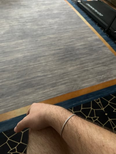 The image shows a close-up view of a person's arm and hand, wearing a thin silver chain bracelet on their wrist. The arm is covered in hair. The person's hand is resting, with fingers curled slightly. The background consists of a carpet with a grey textured pattern, bordered by bands of blue and orange. In the upper right corner, a black piece of equipment, possibly a treadmill, is partially visible. The scene appears to be indoors, possibly in a home or hotel room, and there is no indication of the city of Aqköl, Kazakhstan, or any specific location within it.