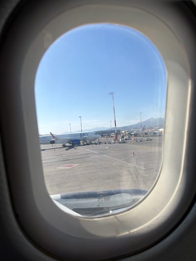 An image taken from inside an airplane shows the view out of an oval-shaped window, looking onto an airport tarmac on a clear, sunny day. The image is rotated 90 degrees counter-clockwise, making the horizon appear vertical.
Through the window, another commercial airplane is visible, parked on the asphalt. Its white fuselage prominently displays "SKY express" in blue lettering. Below this, the phrase "Greece is blu" (likely "Greece is blue") is also visible. The registration number "SX-OBG" can be seen on the aft section of the aircraft. Around the "SKY express" plane, various pieces of ground support equipment, including baggage loaders and service vehicles, are arranged.
In the background, under a bright blue sky, a range of mountains rises, suggesting a mountainous or coastal location. The ground is a light gray tarmac with distinct white and red markings. The interior of the aircraft window frame, which frames the scene, is a light gray color.
