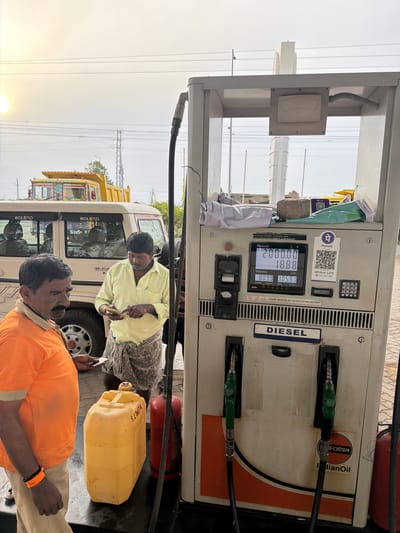 The image depicts a scene at a diesel fuel pump station, likely in Thammarajupalle, India, given the context. In the foreground, two men are interacting near a white diesel pump. The man on the left, wearing an orange shirt and light-colored pants, is standing and holding a small card or receipt. He has a prominent mustache and a dark complexion. To his right, another man, dressed in a light yellow shirt and a patterned lungi, is actively engaged with his mobile phone, holding it in his hands as if performing a transaction or checking information. A large yellow plastic jerrycan is placed on the ground to the left of the fuel pump, with a red fire extinguisher positioned behind it.
The diesel pump itself is white with orange accents and displays a digital screen showing transaction details: "Amount 2000.00", "18.88", "Volume 804.9 Ltr.", and "105.91 Rate / Ltr.". A QR code for PhonePe and a keypad are visible on the right side of the pump. The word "DIESEL" is clearly labeled on the pump. In the background, a beige SUV with "BOLERO" branding is parked. Further back, a bright yellow dump truck with decorative elements on its cabin is visible. Power lines and utility poles suggest an outdoor, potentially semi-rural or roadside, setting. The lighting indicates it is daytime, possibly late afternoon, with a hazy or bright sky. The overall environment suggests a functional fuel station environment.