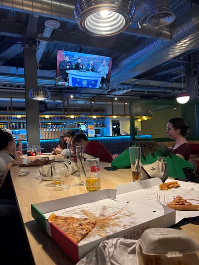 A casual scene depicts a group of people at a high-top table in what appears to be a bar or restaurant. Pizza boxes, some with remaining slices, and multiple empty beer glasses are on the table. A large screen in the background displays a broadcast featuring a panel, potentially a sports show, with an NFL logo present. The setting includes industrial-style exposed ductwork and lighting fixtures, as well as shelves lined with various drinks behind the bar.