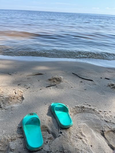 A pair of turquoise slides rests on a sandy beach next to the water. The slides are facing away from the viewer, with the left slide slightly angled towards the camera. The sand is damp and textured, with small ripples and footprints visible. The water is calm with gentle waves lapping at the shore. The sky is visible in the background, with a few wispy clouds. The scene appears to be a peaceful day at the beach in Lyutizh, Ukraine.