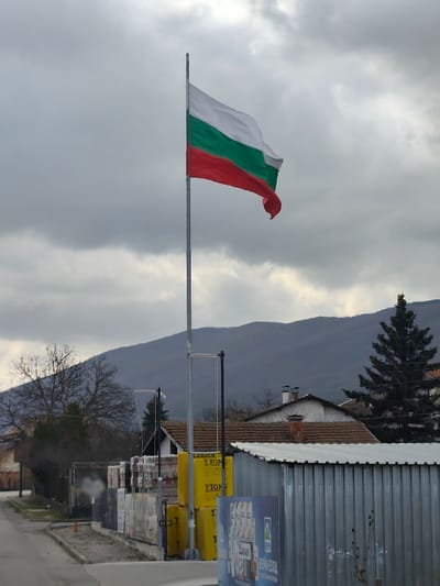 The scene depicts an overcast day in Klenovik, Bulgaria. A tall, silver metallic flagpole dominates the center, flying the Bulgarian national flag horizontally, composed of white, green, and red stripes from top to bottom, waving towards the right under a grey, cloudy sky. In the midground, beneath the flag, several residential structures with brown tiled roofs are visible. One prominent building on the right has a white facade and a chimney emitting no visible smoke. To the right of the flagpole, a stack of yellow construction blocks branded "YTONG" is present, alongside other palletized materials. A corrugated galvanized metal fence or shed runs along the right side of the midground, partially obscuring a blue and white advertisement board displaying Cyrillic text including "ВЕЛИКА ВОДА" and images of bottled water. In the left midground, bare deciduous trees are visible, with a faint plume of white smoke rising among them. A paved road or path is visible in the lower left foreground. Distant mountains with obscured details form the background, their peaks blending with the cloudy sky. No individuals are present in the image.