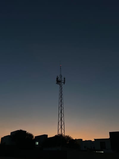 The image depicts a communication tower in Arroyo Hondo, Mexico, silhouetted against a twilight sky. The tall, metallic lattice tower stands centrally, featuring multiple antenna arrays and equipment platforms near its top. A small, red obstruction light is illuminated at the tower's apex. The sky transitions from a dark navy blue at the top to a gradient of orange and peach hues near the horizon, indicating either dawn or dusk. Along the bottom, the dark silhouettes of several buildings and trees are visible. One rectangular window in a building to the left of the tower shows a faint, light-colored internal illumination. No individuals or discernible actions are present in the scene.