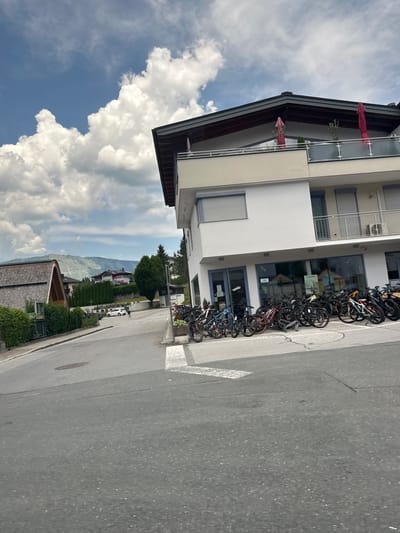 The image depicts a street scene in the city of Au, Austria, on a cloudy day. In the foreground, a grey asphalt road curves to the left. To the right, a modern building with a white facade and a dark-tiled roof is visible. The building has large glass windows on the ground floor, appearing to be a storefront or business. A row of bicycles, likely for rent or sale, is parked in front of the building. A balcony with glass railings and red umbrellas is present on the upper floor of the building.
In the middle ground, a smaller wooden building with a steep, shingled roof is situated to the left. Across the road, a white car is parked. Further back, residential buildings and lush green trees dot the landscape. In the background, rolling mountains form a scenic backdrop under a sky filled with large, fluffy white clouds. The overall impression is of a quiet, picturesque town in a mountainous region.