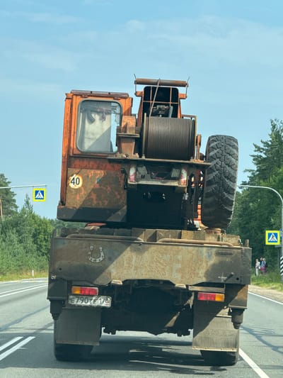 This is a vertically oriented, outdoor photograph taken during the day, likely on a sunny day due to the clear blue sky and shadows cast on the road. The scene depicts the rear and side of a large, orange and rust-colored industrial vehicle, possibly a crane or tow truck, driving on an asphalt road. The vehicle has a large, thick-treaded spare tire mounted on its back. A coiled cable or rope is prominently visible. The cabin of the vehicle is orange with large windows.

On the vehicle's rear, a license plate is visible with the text "E790TE178 RUS" and a horseshoe emblem. A speed limit sign with "40" inside a yellow circle is also affixed to the vehicle's side. In the background, there are trees, indicating a rural or semi-rural setting. A pedestrian crossing sign is visible on a pole by the side of the road. Further down the road, a small group of people, appearing to be two adults and a child, are standing on the grassy verge. The overall impression is of a working vehicle in transit on a road in Russia, with hints of a summer day.