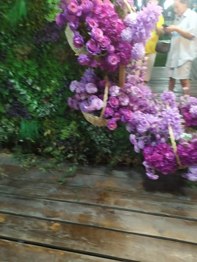 A medium shot captures a rustic wooden platform with an arrangement of artificial purple flowers and greenery. The flowers, in various shades of purple and pink, are displayed in woven baskets and cascade down, some resting on the wooden surface. In the background, a person in a light blue shirt and white shorts is visible, holding a dark object, possibly a phone. Another person in a yellow top and white pants stands nearby. The setting appears to be outdoors, with a hint of green foliage behind the floral display, suggesting a decorative setup in a public space within Sosnovka, Kyrgyzstan. The wooden planks show signs of wear, contributing to a natural, textured environment.