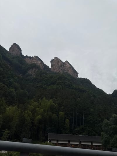 The image depicts a mountain range in the city of Xiehe, China, under an overcast sky. The mountains are partially covered with dense green vegetation. The rock formations are visible in the upper parts of the mountains. A building structure with a dark roof is on the right edge of the frame. A railing is also visible on the right side of the image.