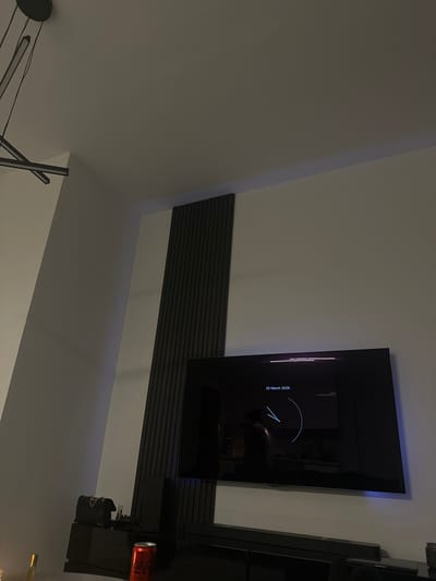 This image displays an interior room located in the city of Latsia, Cyprus. The scene is dimly lit. A large, wall-mounted, flat-screen television is centrally positioned on a light-colored wall. The television screen is active, displaying the text "20 March 2026" and a white circular graphic. Reflections on the screen show the room's interior and a dark, indistinct silhouette of a person facing the television. To the left of the television, a dark, vertical panel composed of parallel slats extends from near the ceiling downwards. Below the television, a dark media console supports a black soundbar. On the console, a black quilted handbag with a metallic chain strap is present on the left, next to a smaller dark object. A red Coca-Cola can is visible further right on the console. The light-colored ceiling features a modern black linear light fixture with multiple arms, suspended in the upper-left portion of the frame. A subtle purple light strip is visible along the top edge of the wall, above the slatted paneling.