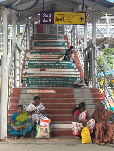 The image shows a wide staircase at a train station. The stairs are decorated with motivational messages in multiple languages, including Tamil, Hindi, and English. The messages focus on cleanliness and environmental awareness, with phrases like "SAVE ENVIRONMENT SAVE NATION" and "CLEAN AND GREEN SHOULD BE OUR AIM." Some steps have small drawings of trees.
At the top of the stairs, a sign indicates "PLATFORM" with the numbers "2 & 3" and "4 & 5." A young man in a striped shirt and light-colored pants is sitting on the stairs, looking at his phone, with a black backpack beside him. Further down, a man in a white shirt and pants is lying down on the stairs, appearing to be sleeping.
On the lower steps, four women are seated. Two women are on the left, one in a yellow and blue sari and the other in a striped white and purple sari, with shopping bags in front of them. Two more women are on the right, one in a white and pink sari with a yellow bag, and another in a brown sari.
The staircase is made of concrete with red tile accents. Metal railings and supports are visible throughout. The background shows a metal roof structure and some greenery, suggesting an outdoor train station environment. The overall scene depicts a busy public space with elements of civic messaging. The presence of multiple languages and the specific cultural motifs on the shopping bags suggest the location is likely in South India, and given the context provided, it is identified as Thanjavur, India.