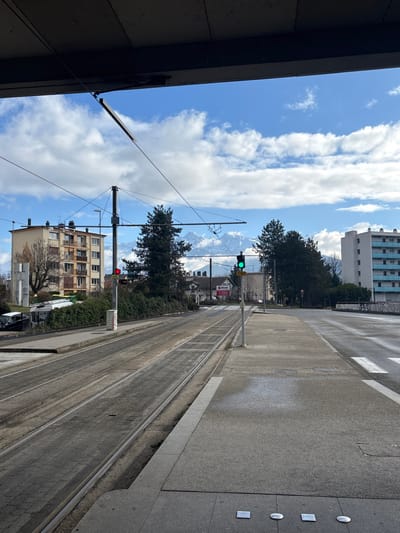 The image depicts a static urban scene in La Tronche, France, captured from underneath a large, dark concrete overpass or elevated structure that spans the top of the frame.

In the foreground, a wide concrete sidewalk with four embedded metallic markers occupies the right side of the frame. To its left, a pair of tram tracks extends into the middle distance, slightly curving to the left, with the ground between the tracks appearing as textured paving. A single green traffic light on a white pole stands on the right side of the tram tracks, displaying a green signal. Further down the tracks on the left, a red traffic light is visible, displaying a red signal. Overhead, a network of black electrical wires, supported by metal poles, crisscrosses the sky, indicative of a tram system.

In the midground, on the left, a multi-story residential building with a beige facade and numerous balconies is visible, fronted by bare deciduous trees and green shrubbery. Directly opposite on the far right, another multi-story residential building with a white facade and light blue balconies is present. Between these buildings, dark green coniferous trees are prominent, partially obscuring smaller structures, including one with a red sign. The tram tracks converge with a paved road surface on the right, where a pedestrian crosswalk is partially visible. Two indistinct figures are visible on the right-hand sidewalk in the distance, walking away from the camera.

The background features a clear blue sky with scattered white cumulus clouds above a distant range of snow-capped mountains, visible beyond the urban landscape and trees. The scene is illuminated by bright daylight, casting subtle shadows. The overall environment appears orderly and well-maintained.