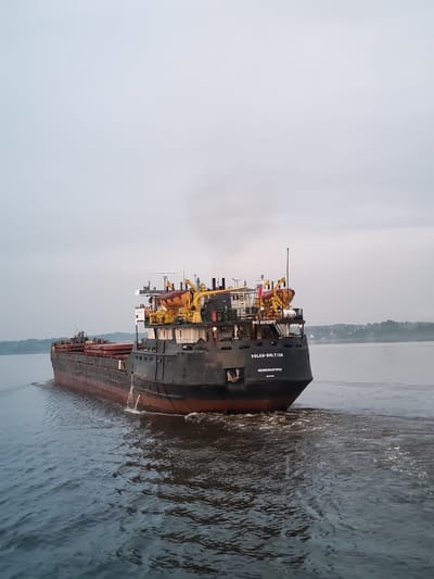 A cargo ship named "VOLGO-BALT 136" is seen from its stern, sailing on a body of water. The ship is dark gray with a red waterline. It has a superstructure with yellow lifeboats and various antennas. The name of the ship is clearly visible in white lettering on the stern, along with smaller text below. Smoke is emanating from a stack on the ship. The water is choppy and a wake is visible behind the vessel. The background shows a hazy shoreline with trees and some buildings, suggesting a temperate climate under an overcast sky. The location is identified as the city of Ratibor, Russia, though the image itself does not contain specific landmarks that confirm this location, other than the name on the ship which may be indicative of its operational area. There are no people visible in the image.