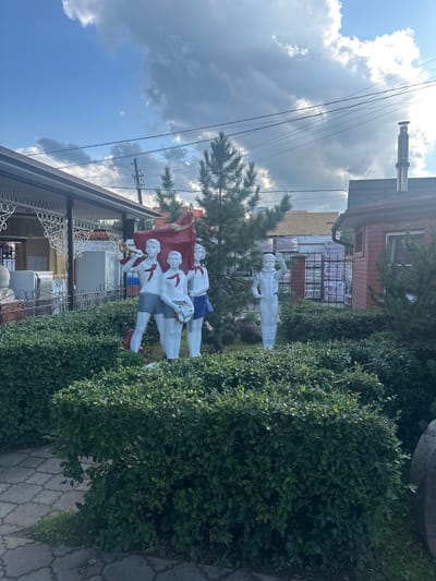 In Kablukovo, Russia, a group of four white statues stands in front of a meticulously trimmed green hedge. The statues depict young people, possibly students, dressed in uniforms. Three of them are grouped together; one plays a trumpet, another holds a flag that appears to be red, and the third clutches a book or notebook. The fourth statue, positioned slightly apart, stands dressed in a spacewalk suit, with one arm raised in a wave. Behind the hedge, a paved walkway is visible, along with a glimpse of building structures, including a brick building with a chimney and a covered patio. The sky above is a mix of blue and dramatic, fast-moving clouds, suggesting dynamic weather.