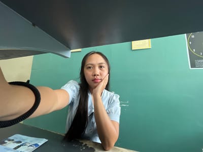 The image shows a young woman taking a selfie. She is wearing a light blue collared shirt with puffed sleeves and has long, dark hair. She is resting her chin on her hand, looking towards the camera with a neutral expression. She has a black coiled bracelet on her left wrist. The background is a teal-green wall with two framed certificates and a target poster with numbers from 7 to 10. A dark, angled structure is visible in the foreground, partially obscuring the top of the frame. The scene appears to be indoors, possibly in an office or a public building. The presence of a target poster suggests a possible connection to a shooting range or a similar establishment. The certificates on the wall might indicate awards or official recognitions. The overall lighting suggests an indoor setting, likely during the day, with natural light contributing to the scene.