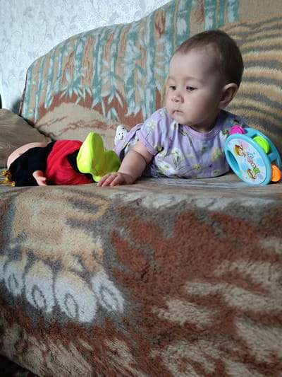 In Kamennoye, Russia, a baby lies on a couch with a doll and a toy. The baby, facing right, wears a lavender shirt with a floral pattern. A doll, dressed in red and black with yellow accents, is positioned to the baby's left. A blue tambourine toy, featuring colorful images, sits to the baby's right. The couch is covered with a patterned blanket.