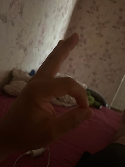 The image shows a person's hand making an "okay" gesture in the foreground. The hand is positioned in the center-right of the frame, with the thumb and index finger forming a circle. The background is out of focus and dimly lit, showing what appears to be a bed with pink bedding and pillows. A white power adapter with a cable is visible on the bedding near the bottom of the frame. The wall behind the bed is covered in wallpaper with a floral pattern. The lighting suggests an indoor setting, possibly a bedroom, and the overall atmosphere is casual and relaxed. There is no information in the image that provides any context or details about the city of Saran, Kazakhstan.