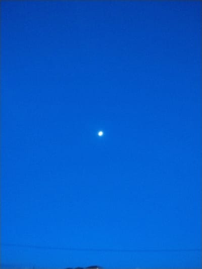 A vibrant blue sky dominates the image, fading to a slightly darker hue near the top. A single, bright white point of light, presumably a star or the moon, is centered in the composition. At the bottom of the frame, a faint horizontal line suggests either wires or a horizon. Below this line, a barely discernible darker shape hints at the presence of buildings or vegetation.