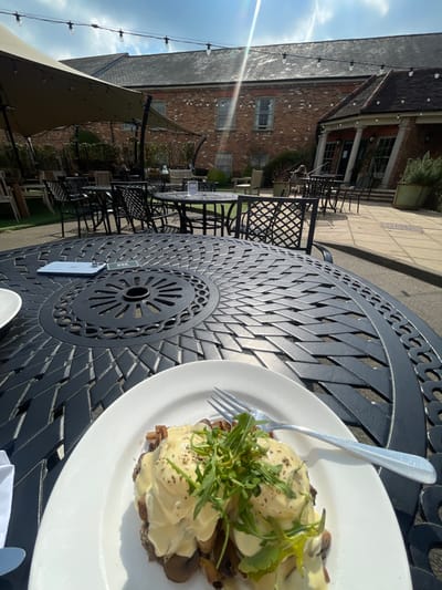 The image shows a plate of food, a white plate with two poached eggs with hollandaise sauce and mushrooms, on top of a black metal table with a circular pattern. There are other black metal chairs and tables around the table, as well as a white table with a blue phone and a white object on it.  The background shows a brick building with a tan awning over a patio area with a light green grassy area between the building and the table. There are some trees and bushes visible. It is a sunny day, and the light shines through the awning. The location is at latitude 51.989, longitude -0.619, but this location does not correspond to a recognizable place in Woburn, United Kingdom. The coordinates may be incorrect or Woburn may not have an address that corresponds to the image location.