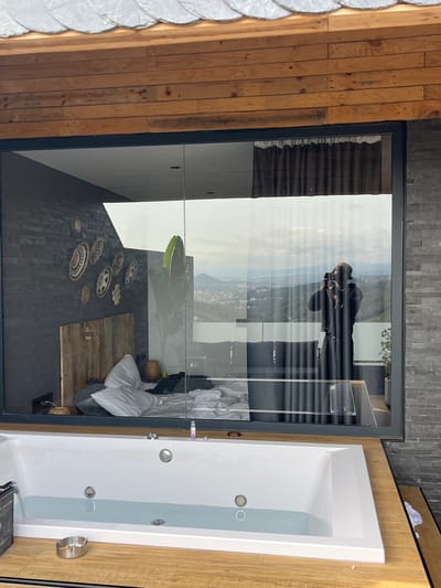The image shows a hotel room with a large window overlooking a mountainous landscape. The room features a bathtub filled with water in the foreground, a bed with pillows, and a wall decorated with woven circular items. In the reflection of the window, a person wearing a hat and jacket is visible, holding a phone and appearing to take a photo. The exterior environment appears to be a city or town nestled in the mountains. The architecture suggests a modern design with wood and stone elements. The room is lit by natural light.