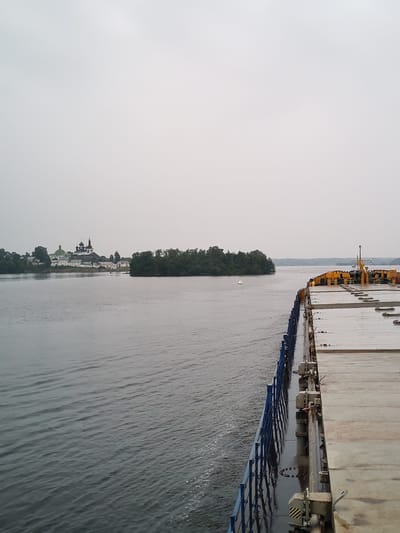 The image captures a water view from the deck of a barge or ship, looking towards land on the left. On the distant left bank, a monastery complex with white buildings and green and black domed roofs is visible, set against a backdrop of trees. The monastery is identified as the Goritsy Monastery of Resurrection, located in Goritsy, Russia, on the shores of the White Lake. A small green islet with trees is situated in the middle of the water in front of the monastery. The foreground is dominated by the deck of the vessel, featuring a flat, grey surface with visible metal cleats and a blue railing along the edge. Yellow cranes or equipment are also present on the deck. The water is a calm, grey expanse, with small ripples. The sky is overcast and a pale grey. There are no people visible in the image.