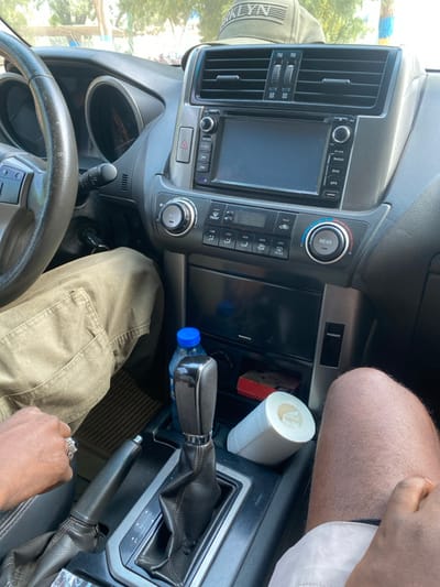 The image depicts the interior of a vehicle, specifically the dashboard, central console, and portions of the driver and front passenger, located in Yan Dala, Nigeria.

In the driver's seat, a person wears khaki-colored cargo pants, with their left hand, adorned with a ring on the index finger, resting near the handbrake lever. The steering wheel is partially visible on the left. Keys are in the ignition.

The central console features an infotainment system with a dark screen, flanked by air vents and various buttons for climate control, hazard lights, and media functions. Below the infotainment system, an automatic gear shifter is visible. To the right of the gear shifter, a blue plastic water bottle stands upright. Adjacent to the bottle, a small red rectangular object, possibly a box of matches, and a white roll of paper, resembling toilet paper or a paper towel roll, are present.

In the front passenger seat, the upper part of a person's bare right thigh is visible, indicating they are wearing light-colored shorts or undergarments. Above the dashboard, a green baseball cap with "BROOKLYN" embroidered on it rests. The scene is illuminated by natural daylight entering through the windshield.