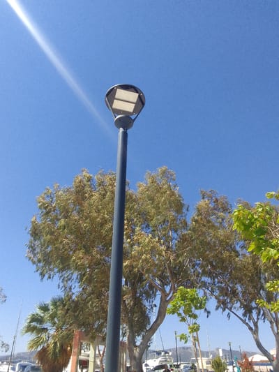 A tall, dark gray lamp post stands in the foreground against a bright blue sky, topped with a circular fixture containing two rectangular panels. Sunlight streams in from the upper left, creating a bright ray across the sky. Below the lamp post, lush green and brown foliage from several trees fills the middle ground, with a palm tree visible to the left. In the background, a body of water with boats and yachts is visible, along with some buildings and a distant hilly landscape. The scene appears to be a daytime shot in a coastal area, possibly a marina or promenade, located in the Municipal Unit of Elefsina, Greece.