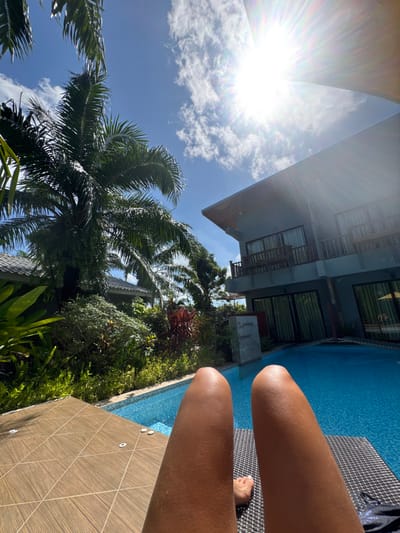 Sunlight streams through a sky with scattered clouds, illuminating palm trees and tropical foliage near a building. A blue pool borders the scene. The tanned legs of a person sitting beside the pool are visible. The surrounding area appears lush, suggesting a tropical vacation setting.