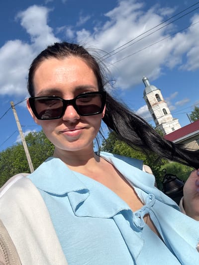 An outdoor selfie captures a woman from the chest up under a bright blue sky with scattered white clouds, suggesting a clear and sunny day. The woman wears dark, rectangular sunglasses that obscure her eyes, and her lips show a slight, neutral smile. Her dark hair is pulled back. She is dressed in a light blue top featuring ruffles and a decorative knot tied at the neckline, possibly over a lighter, white or beige garment.
In the background, to the right of the woman, an old, light-colored architectural building, resembling a church or bell tower with distinct windows and a dome-like roof, stands against the sky. Green trees are visible in the mid-ground. Thin power lines stretch across the upper left portion of the sky, and a wooden utility pole is present in the lower background. The scene suggests a rural or small-town setting, fitting the provided context of Луптюг, Russia. No visible text is present in the image.