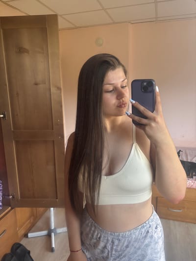 A young woman is taking a selfie in a mirror. She is wearing a white bralette and patterned shorts. Her hair is long and dark brown. She is holding a dark purple phone in her right hand. Her nails are painted with a white and blue design. The setting appears to be a bedroom, with a wooden dresser in the background. The lighting suggests it is daytime. The woman has a slight smile on her face. There is no visible text in the image.