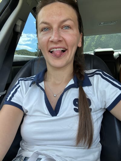 A woman is pictured in the driver's seat of a vehicle, possibly a car, with a black leather interior. The woman is facing the camera, and her head is slightly tilted. She has blue eyes, light brown hair braided into a single plait, and is wearing a white polo shirt with navy blue trim and a diamond pendant necklace. Her tongue is sticking out. The background shows a glimpse of a blue sky with clouds, green hills, and trees, suggesting an outdoor setting. The image does not provide any information to place the location in Dulovine, Montenegro.